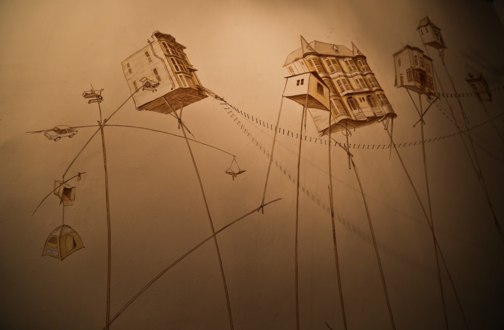 "The Mission Condition - Outwardly Mobile" Mural and Assemblage by Mona Caron and Dustin Fosnot, depicting San Francisco houses teetering precariously on stilts surrounded by alternative housing such as tents, cardboard boxes and shopping carts. Photo by Isabeau Doucet